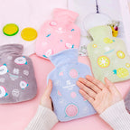 Quatre bouillottes peluche bleue, rose, grise et verte disposées sur fond rose, mains posées sur la bleue